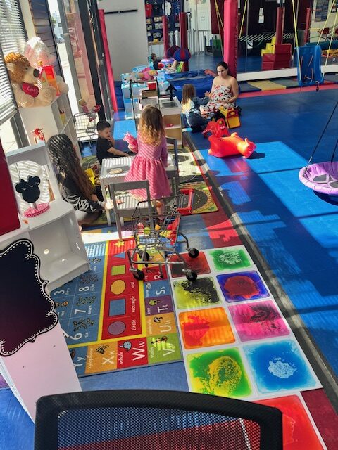Children enjoying various play activities in a colorful indoor playground. A girl in a pink dress stands near a mini grocery cart, while other children engage with toys and play equipment in a bright, inviting space filled with educational and interactive elements.